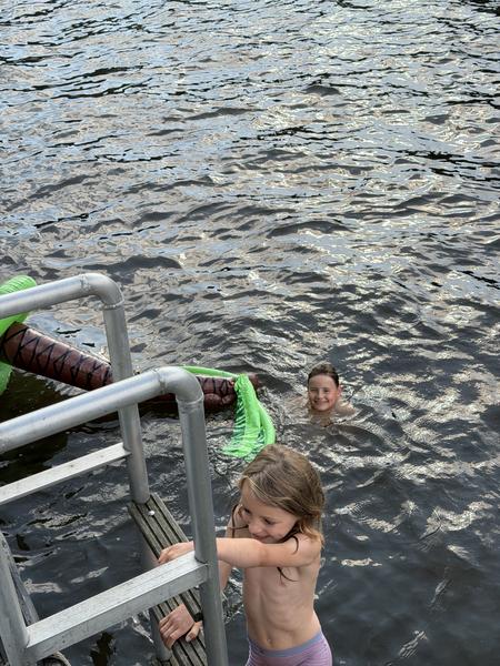 Two children are enjoying a swim, with one climbing a ladder and the other smiling from the water, next to an inflatable palm tree